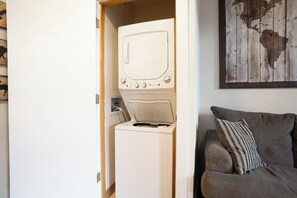 A compact room with a washing machine and dryer, a gray couch with a striped pillow, and a wooden wall with a map.