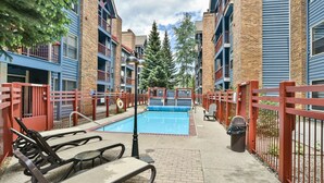 A pool area with lounge chairs and a trash bin, surrounded by multi-story buildings with balconies and a large tree in the center. - River Mountain Lodge Aspen Trail Condo: Ski In, Downtown Breck (Breckenridge)