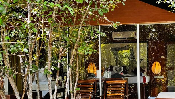 Ένας εξωτερικός χώρος με καθίσματα, με ξύλινα τραπέζια και καρέκλες, που περιβάλλεται από δέντρα και διαθέτει ένα γυάλινο περίβλημα.