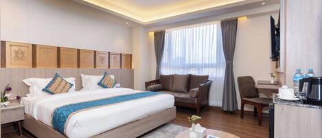A hotel room with a large bed, a sitting area with a chair and a table, a desk with a computer, and a window with curtains.