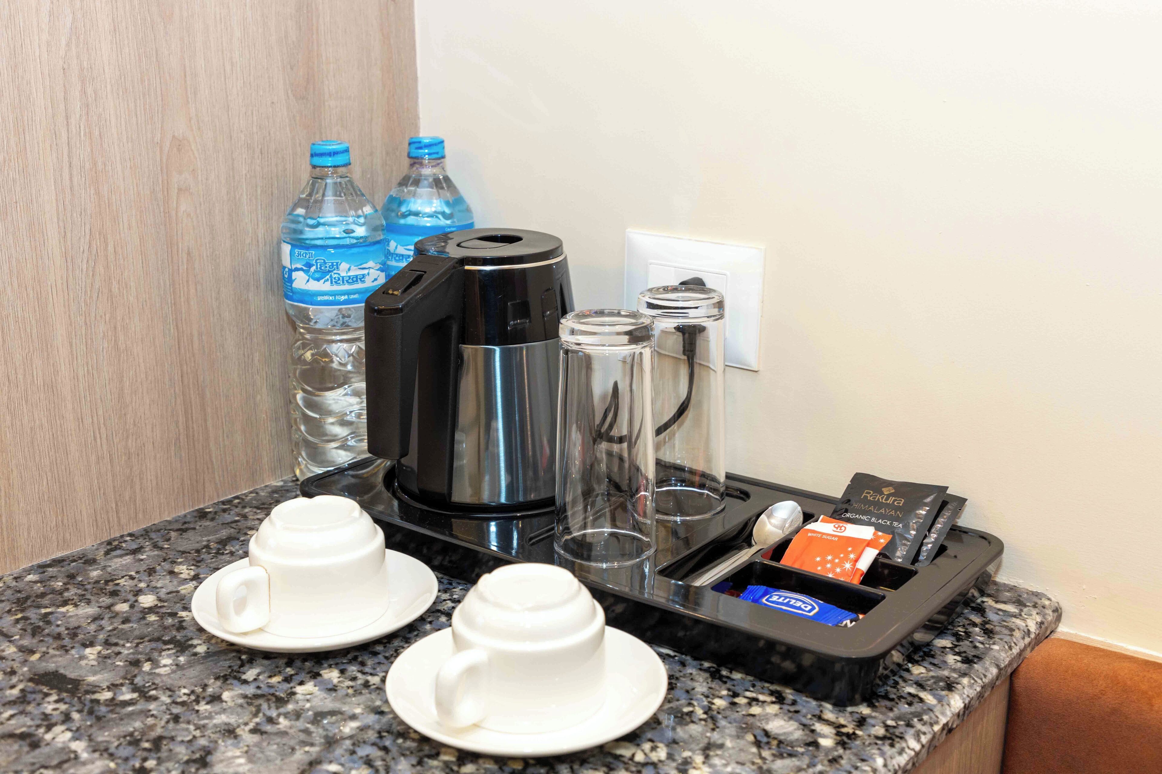 A coffee maker, two cups, two glasses, and a tray with tea and coffee pods on a granite countertop.