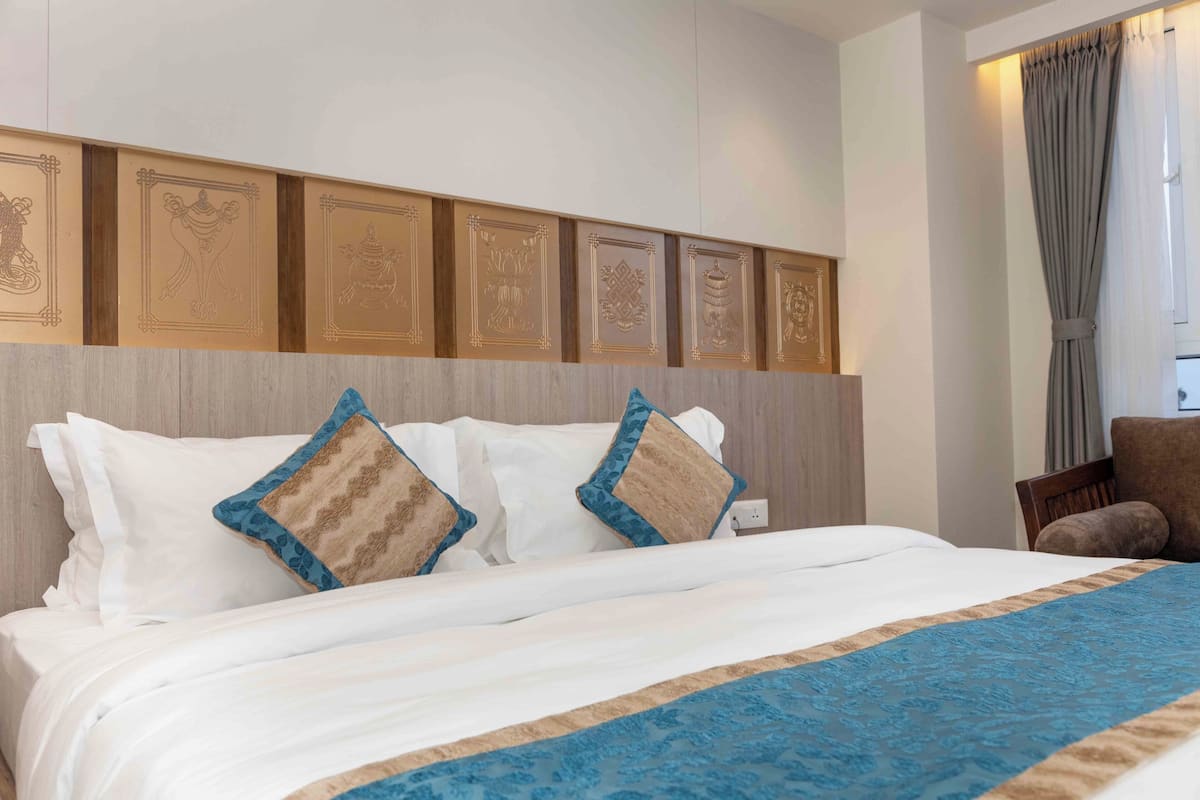 A hotel room with a bed, pillows, a headboard with decorative panels, a chair, and a window with curtains.
