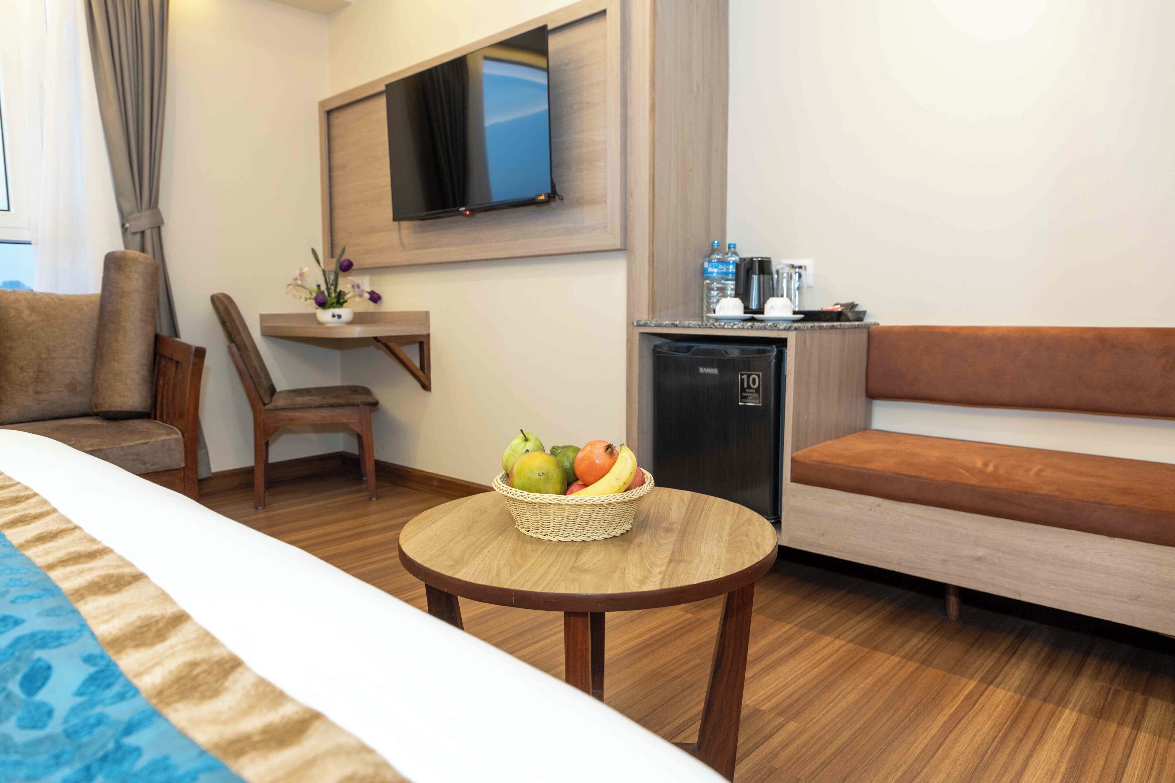 Una habitación de hotel con una cama, una mesa de madera con una cesta de frutas, un sofá, una silla, un televisor montado en la pared y un minibar.