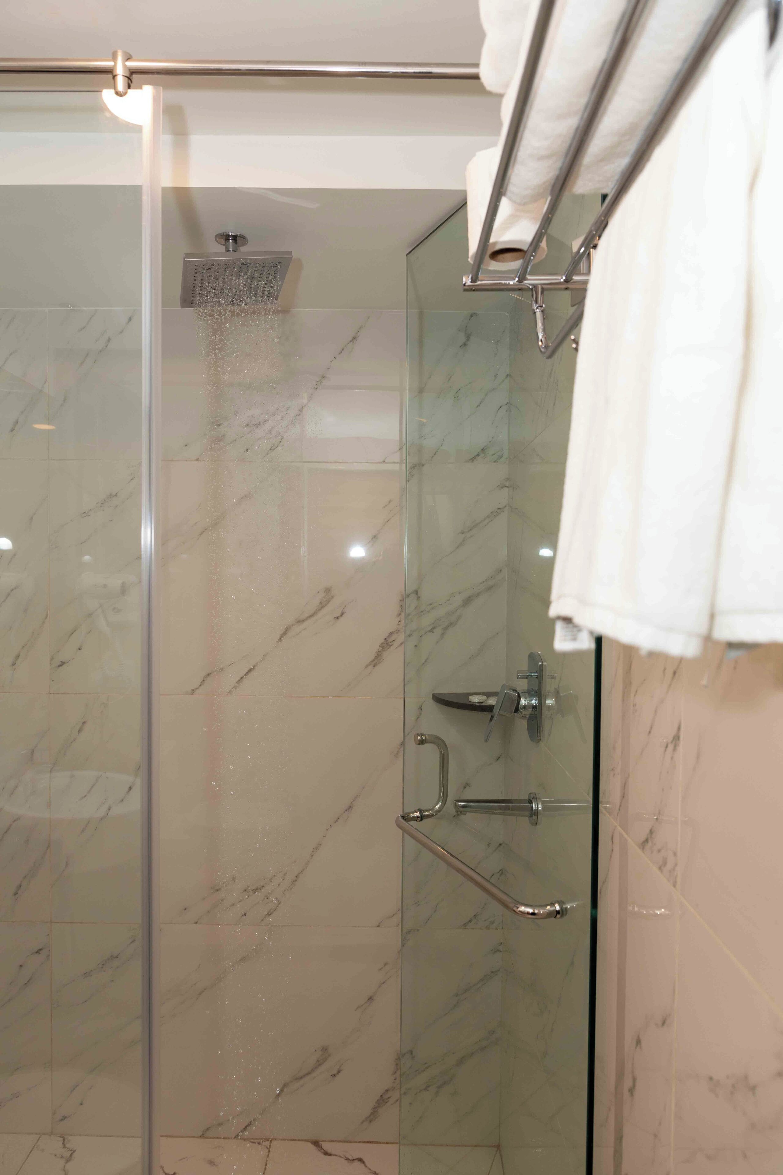 A glass-enclosed shower with a handheld showerhead, a handheld shower control, and a towel rack with a white towel.