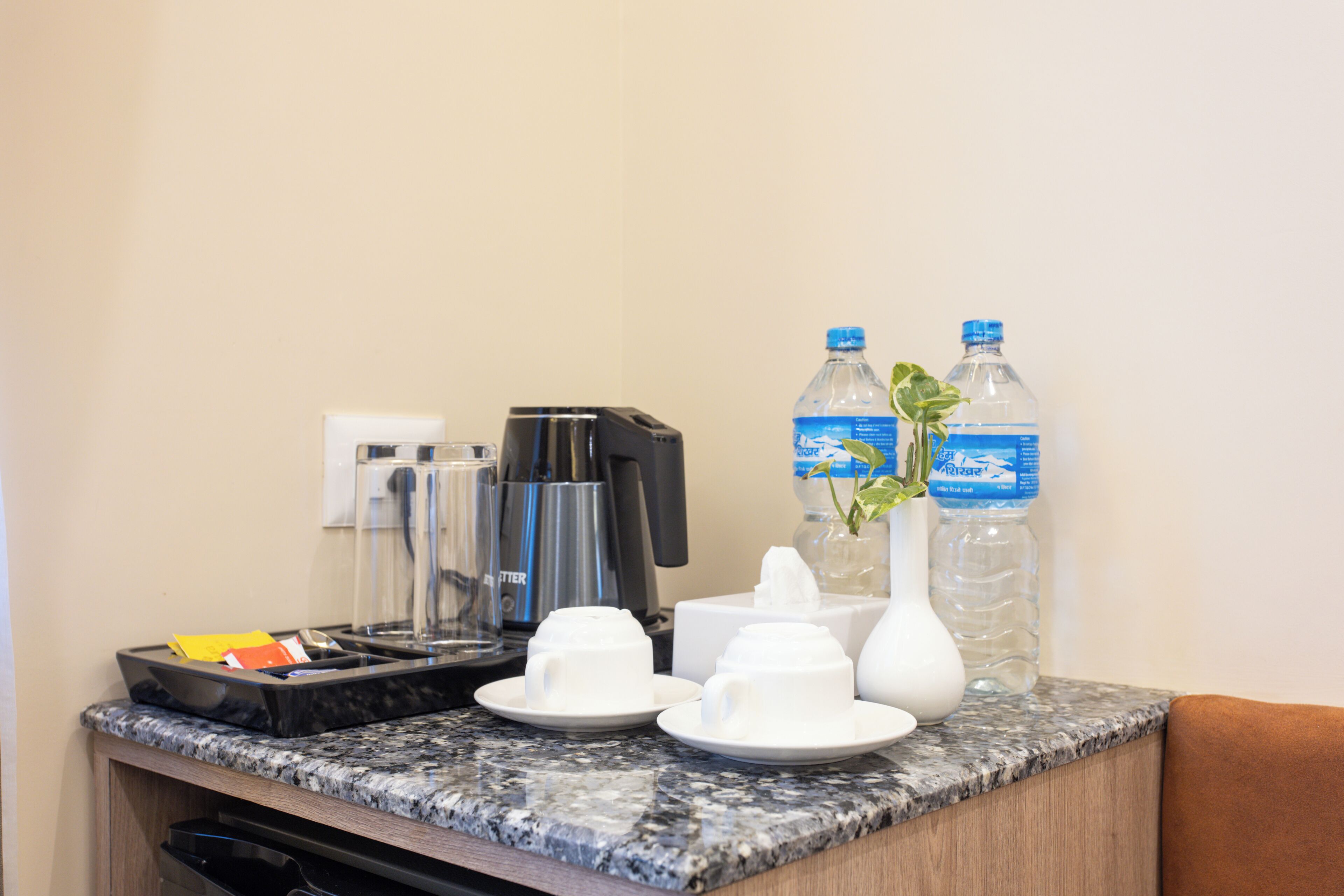 A hotel room's breakfast bar with a coffee maker, two water bottles, a vase with a plant, and a trash bin.