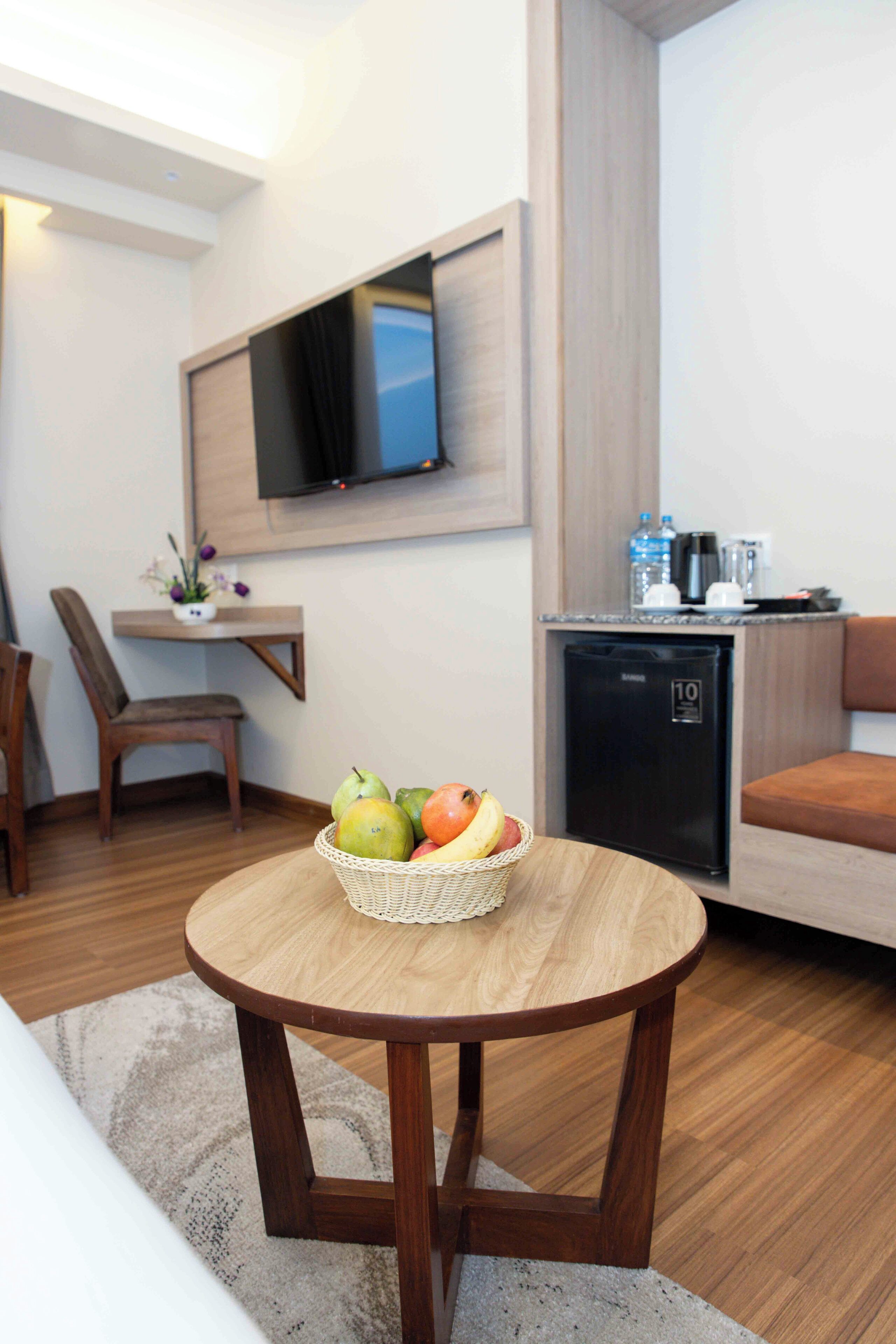 A modern hotel room with a wooden dining table, a small kitchen area with a refrigerator, a flat-screen TV mounted on the wall, and a comfortable seating area.