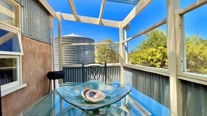 A glass table with a decorative bowl and a chair in a sunlit room with large windows and a view of trees and a water tower. - Self contained cabin in quiet coastal bushland setting. Pet friendly! (Robe)
