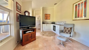 A living room with a TV, wooden entertainment unit, grey armchair, and a kitchen in the background. - Self contained cabin in quiet coastal bushland setting. Pet friendly! (Robe)