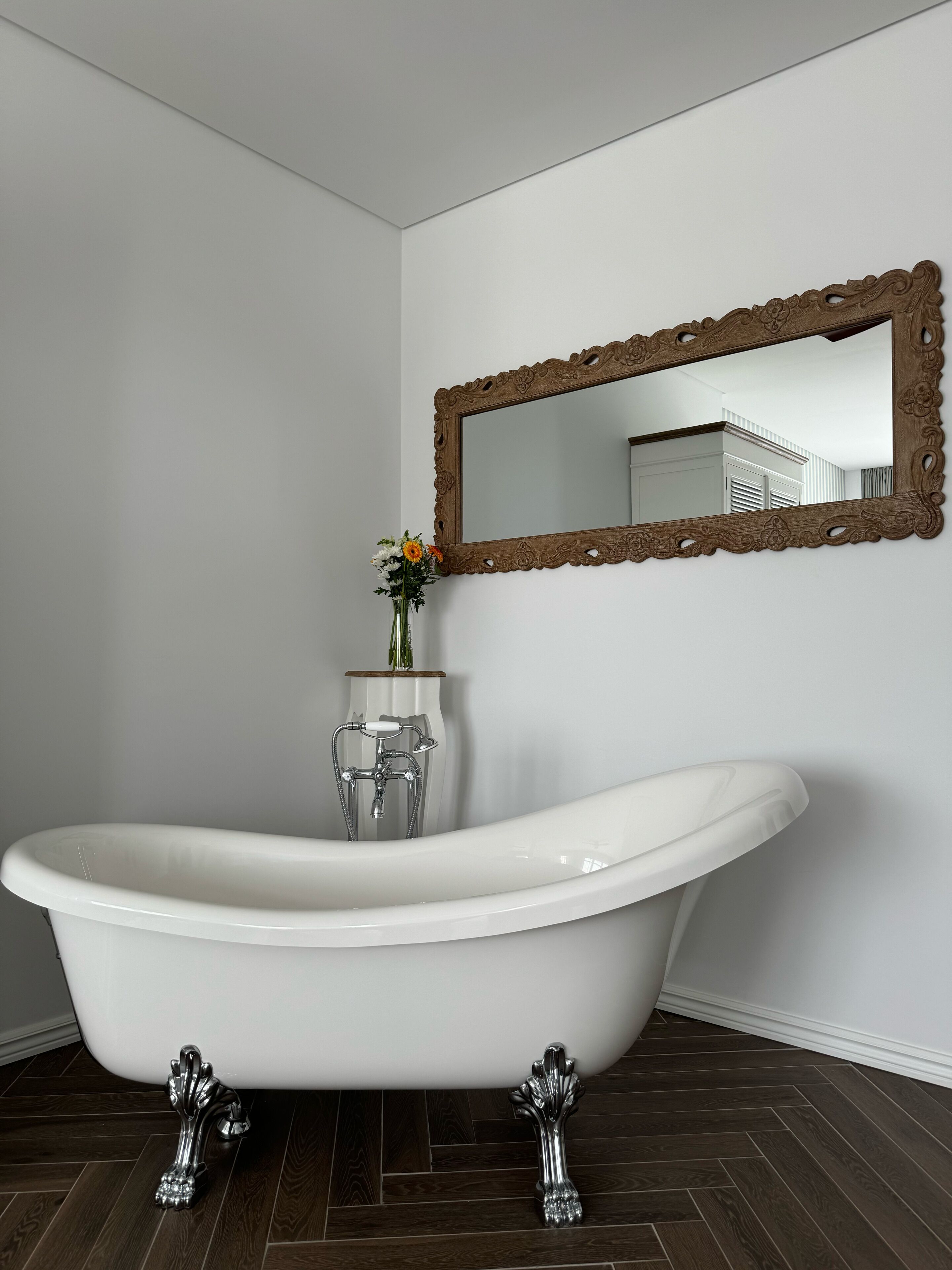 A bathroom with a freestanding bathtub, a wooden-framed mirror, and a vase with flowers on the wall.