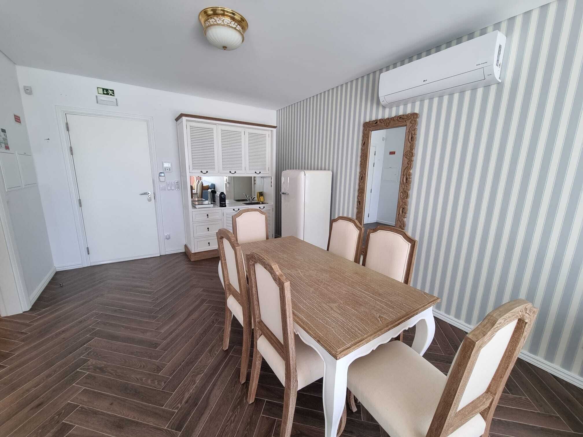 A dining area with a wooden table and chairs, a kitchenette with white cabinets, and a striped wallpaper wall.
