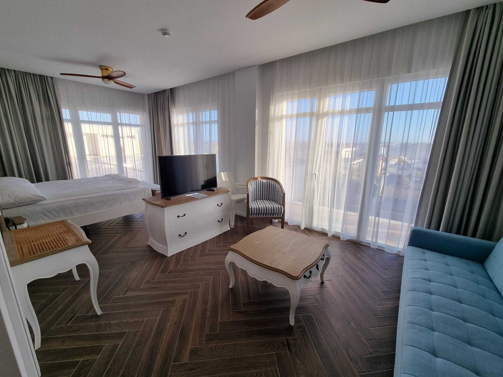 A hotel room with a bed, a TV on a dresser, a chair, a sofa, and large windows with curtains.