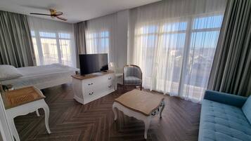 A hotel room with a bed, a TV on a dresser, a chair, a sofa, and large windows with curtains.