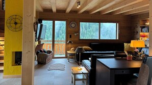 A cozy cabin interior with a wooden ceiling, a fireplace, a sofa, a dining table, and a bookshelf. - New luxury chalet in Morzine's La Cote d'Arbroz with a Jacuzzi, beautiful views, and a quiet setting (La Côte-d'Arbroz)