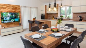 A modern dining area with a wooden table set for six, a large flat-screen TV on the wall, and a kitchen in the background.