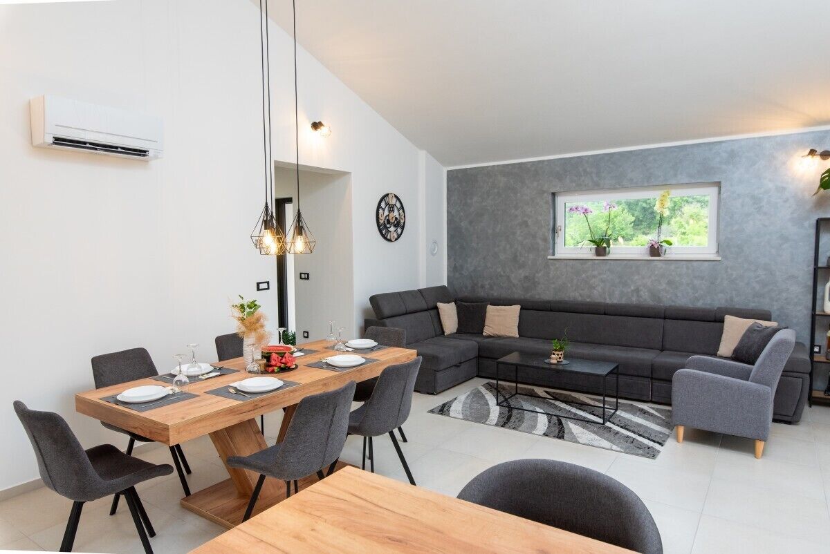 A modern living room with a dark gray sectional sofa, a wooden dining table with chairs, and a large window with plants.