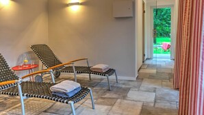 Een wellnessruimte met twee zwart-wit gestreepte lounge fauteuils, een klein rood tafeltje met een glas, en uitzicht op een groen buitengebied door een glazen deur.