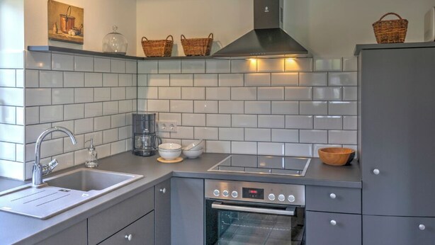 Een moderne keuken met grijze kastjes, een roestvrijstalen spoelbak, een ingebouwde oven en een afzuigkap. Op het aanrecht staan twee rieten manden en aan de muur hangt een schilderij.