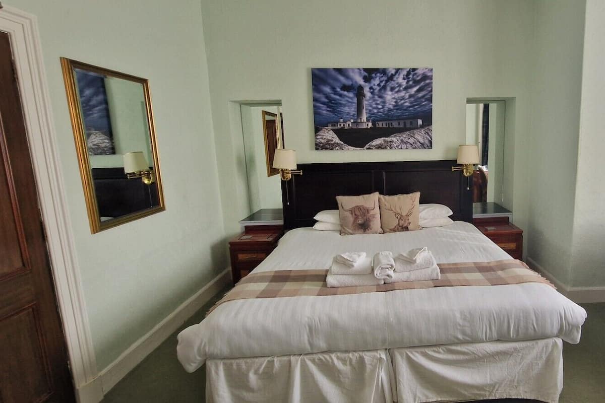 A hotel room with a bed, bedside tables, a desk, a chair, a mirror, and artwork on the wall.