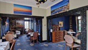 A dining area with tables and chairs, a fireplace, and a view of the sea through a window. - Corsewall Lighthouse Hotel (Stranraer)