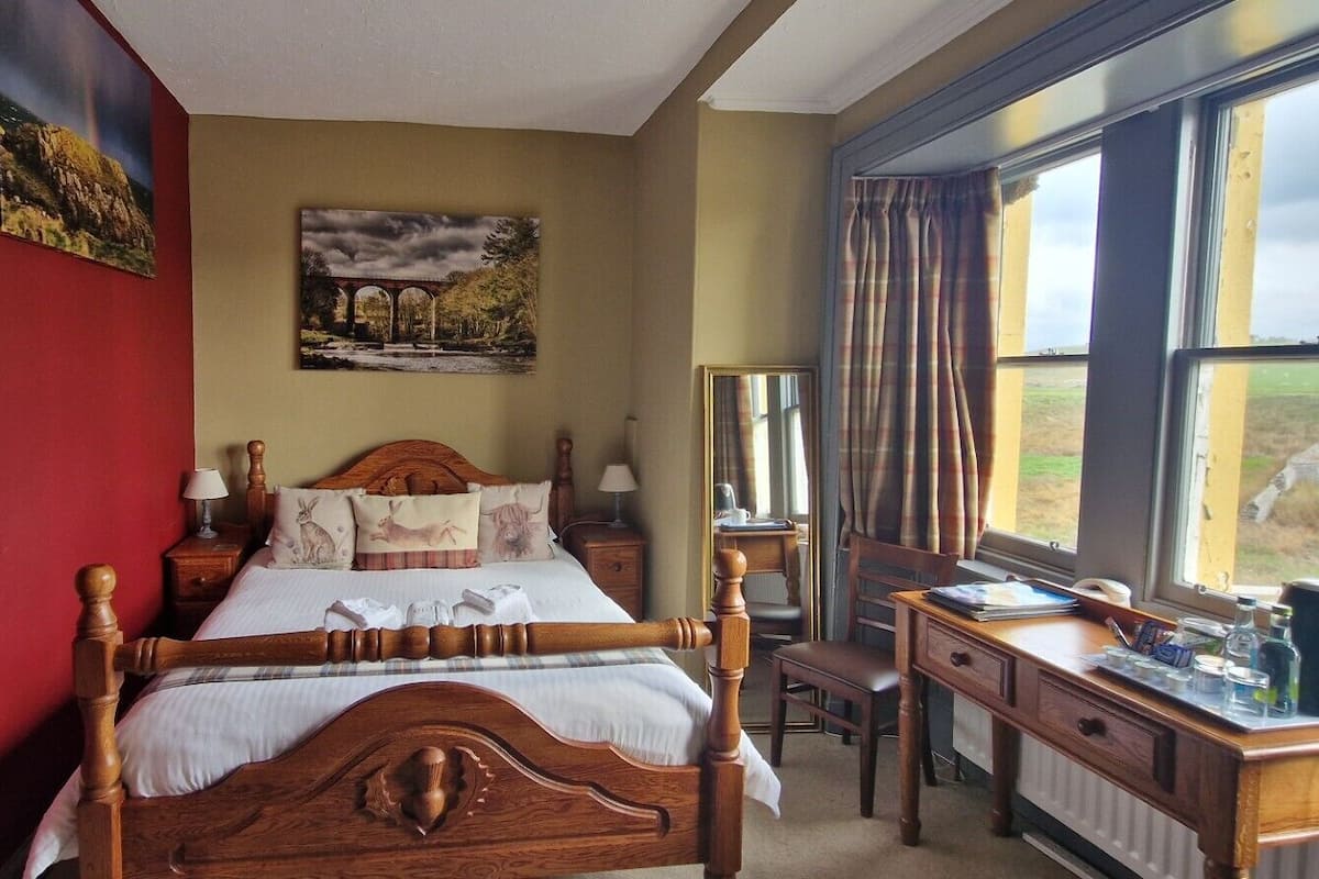 A bedroom with a wooden bed, a desk, and a view of the countryside through large windows.