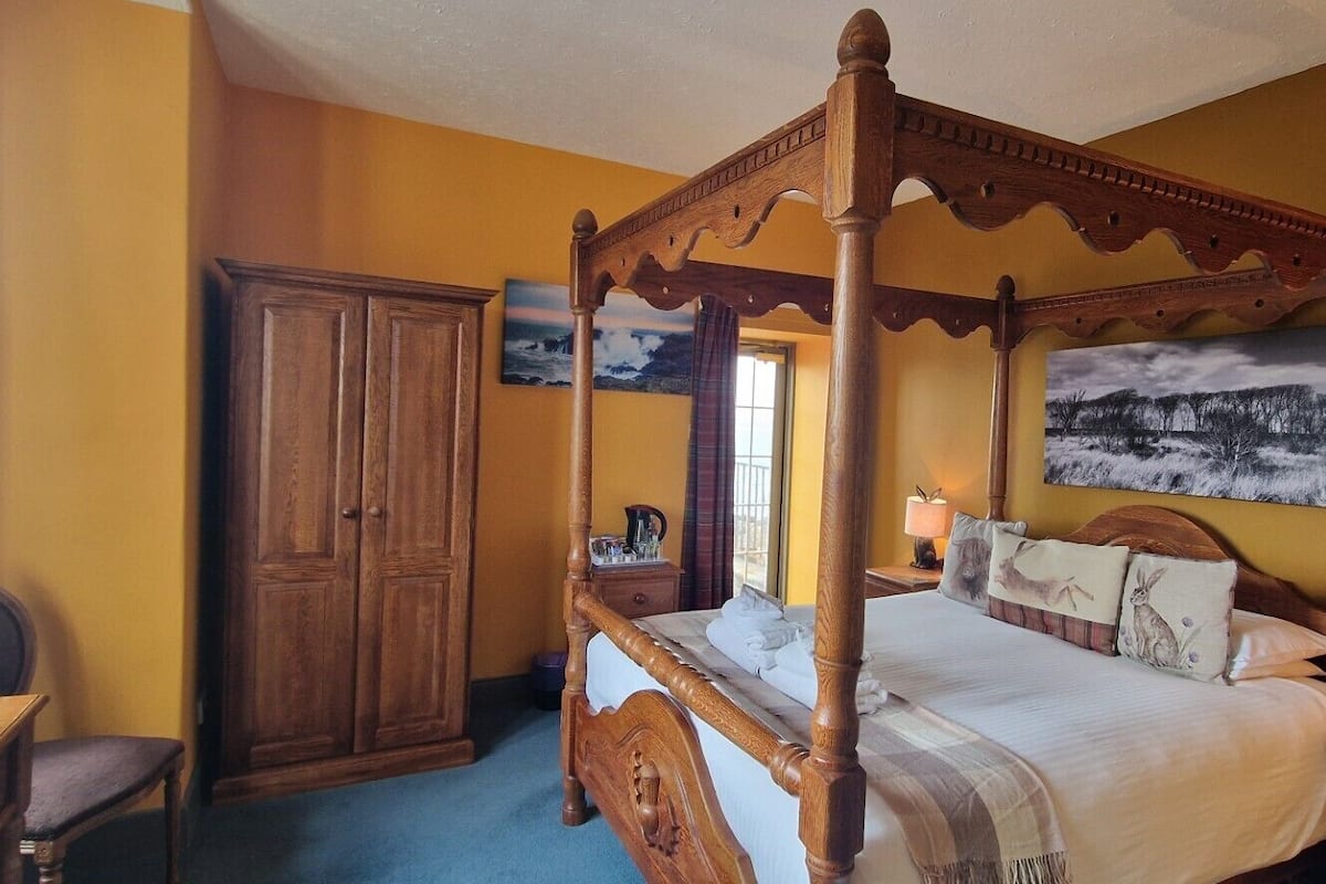 A four-poster bed with a wooden frame, a wardrobe, a desk with a chair, and a painting on the wall.
