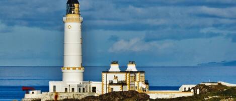 Un faro con una torre blanca y un edificio más pequeño con detalles amarillos, ubicado en una costa rocosa con el mar al fondo.