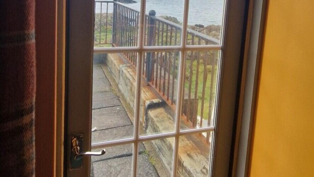 A view through a window with a metal grid, overlooking a paved area with a railing and a grassy field. - Corsewall Lighthouse Hotel (Stranraer)