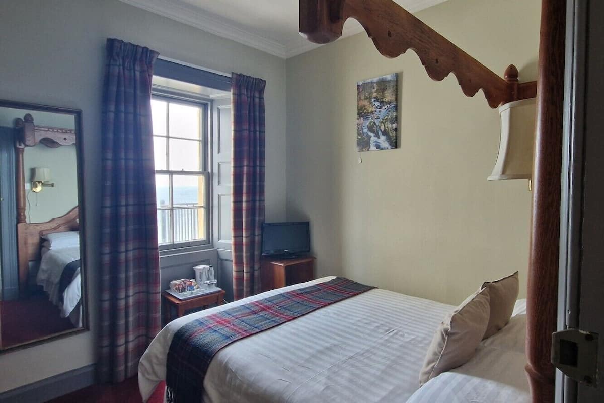 A hotel room with a bed, a mirror, a window with curtains, a painting on the wall, and a wooden ceiling beam.