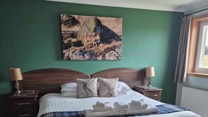 A neatly made bed with a plaid blanket and two pillows, a painting of a deer on the wall, and a window with curtains. - Corsewall Lighthouse Hotel (Stranraer)