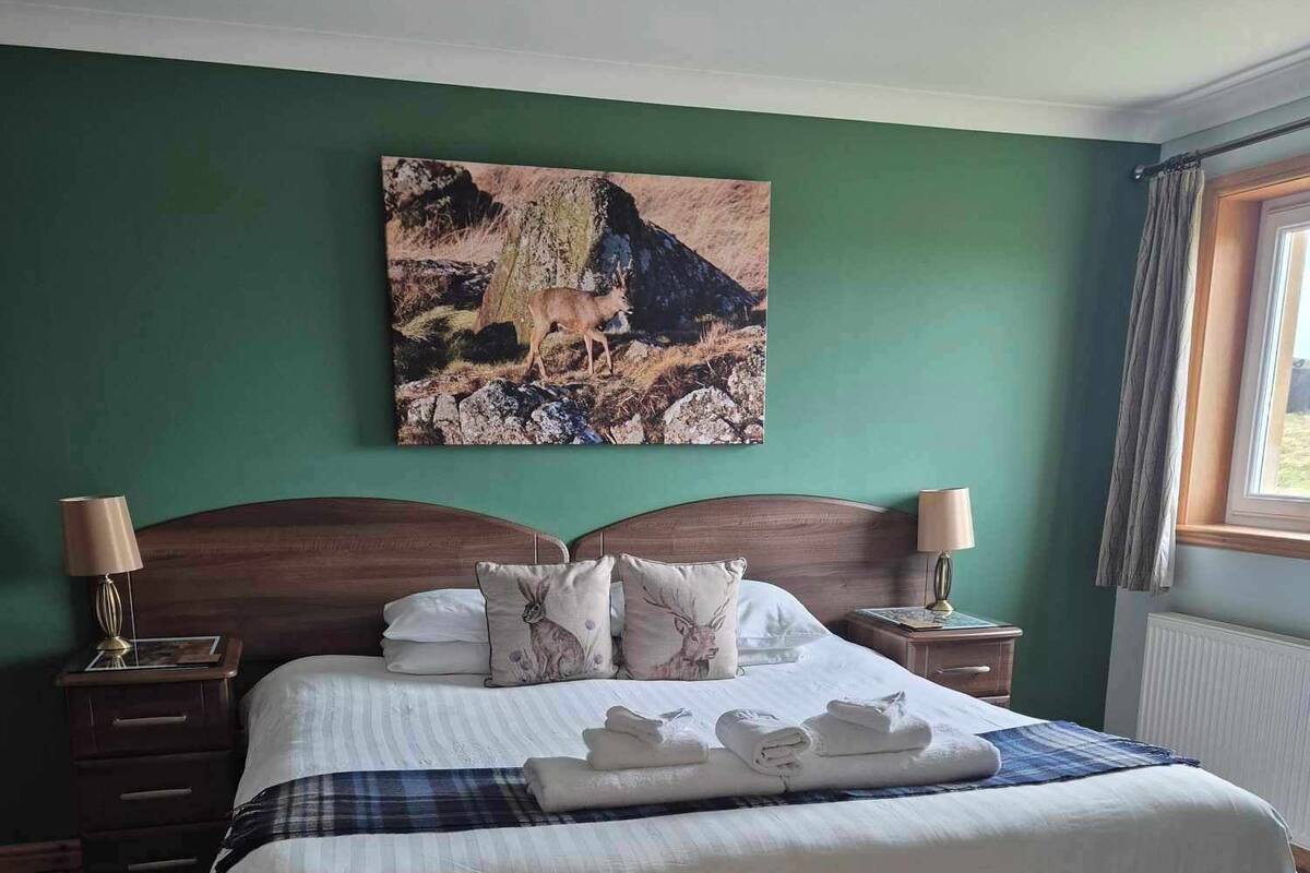 A neatly made bed with a plaid blanket and two pillows, a painting of a deer on the wall, and a window with curtains.