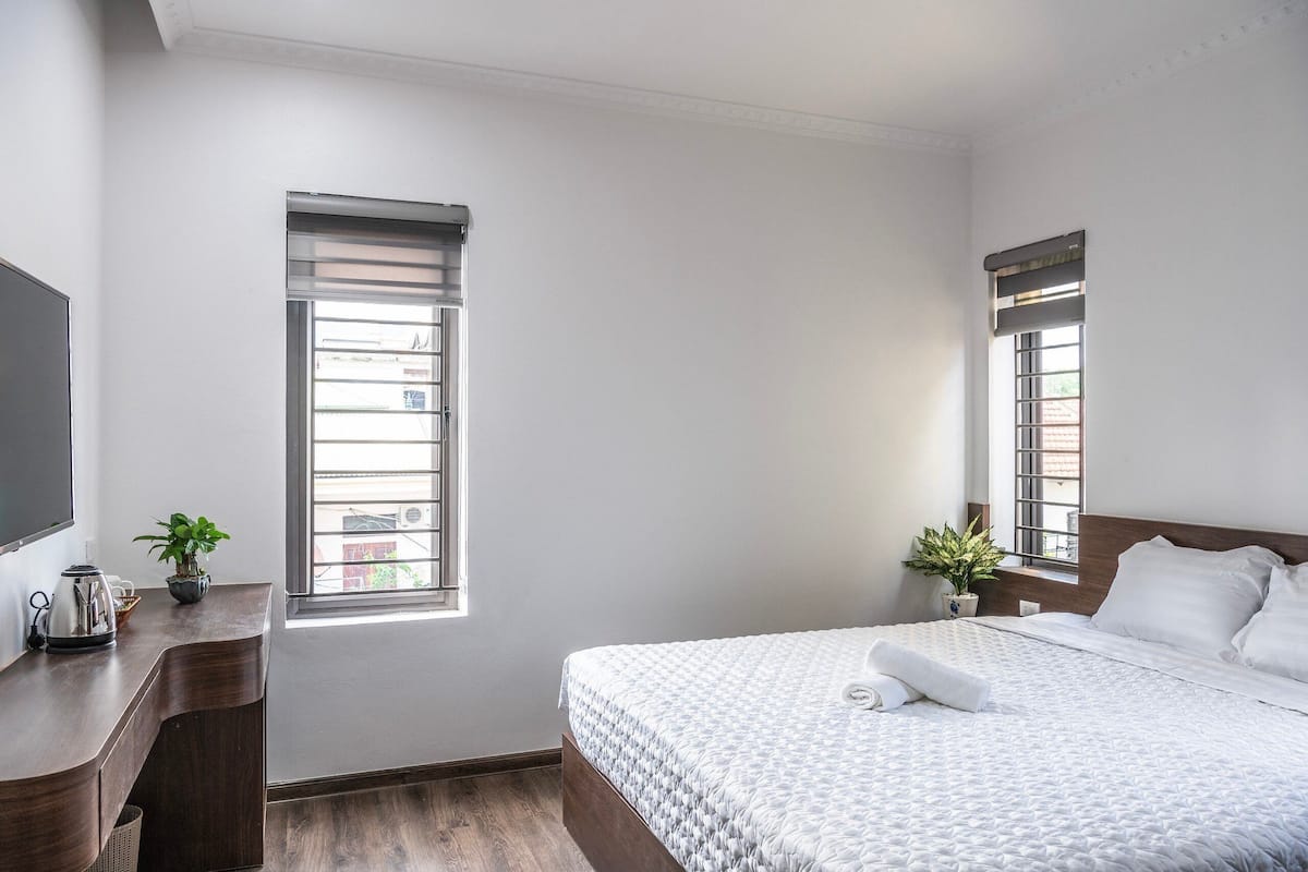 A bedroom with a bed, a desk, a potted plant, and a window.