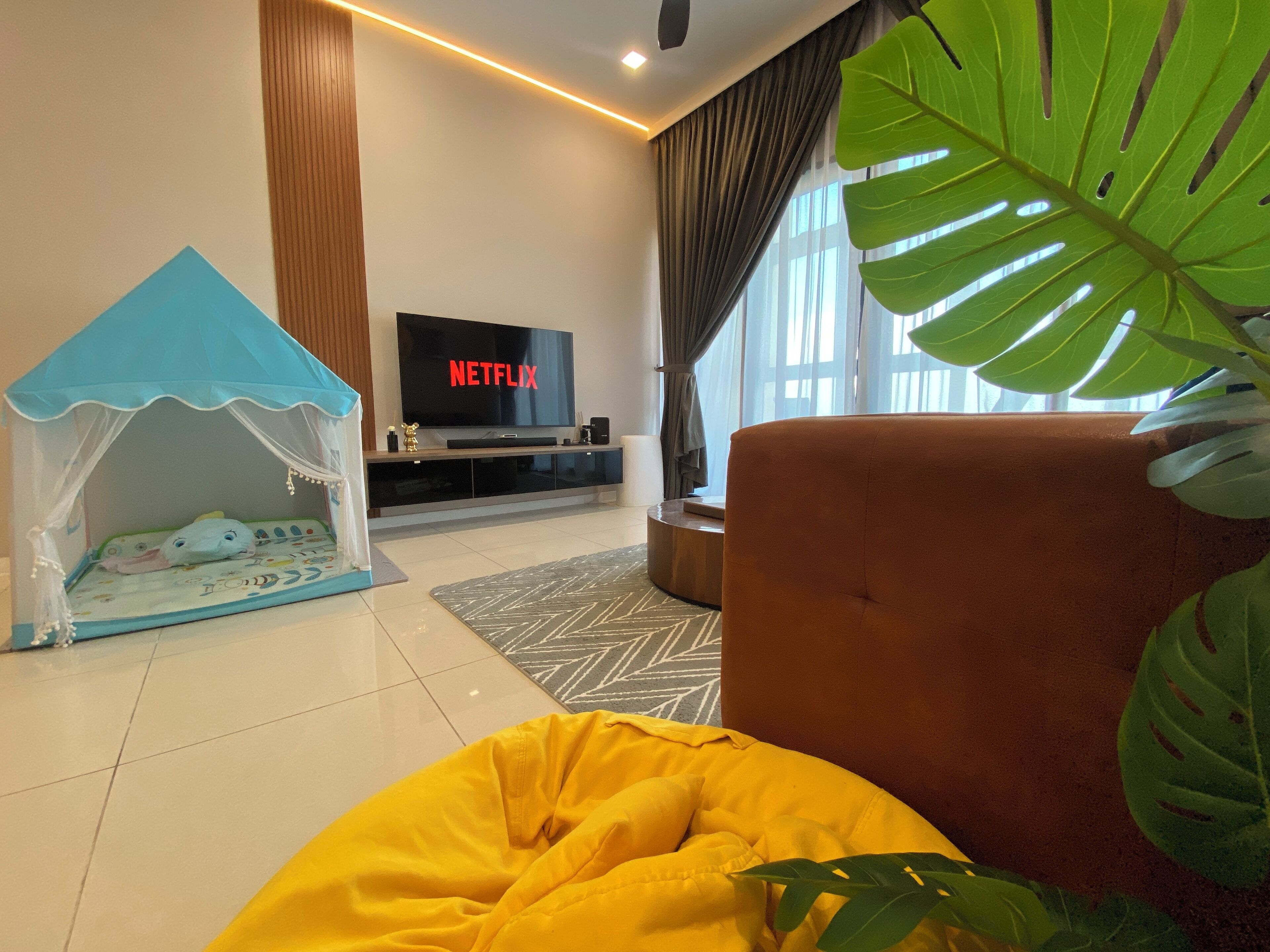 A living room with a TV displaying 'NETFLIX', a playpen, a yellow bean bag, and a large plant.