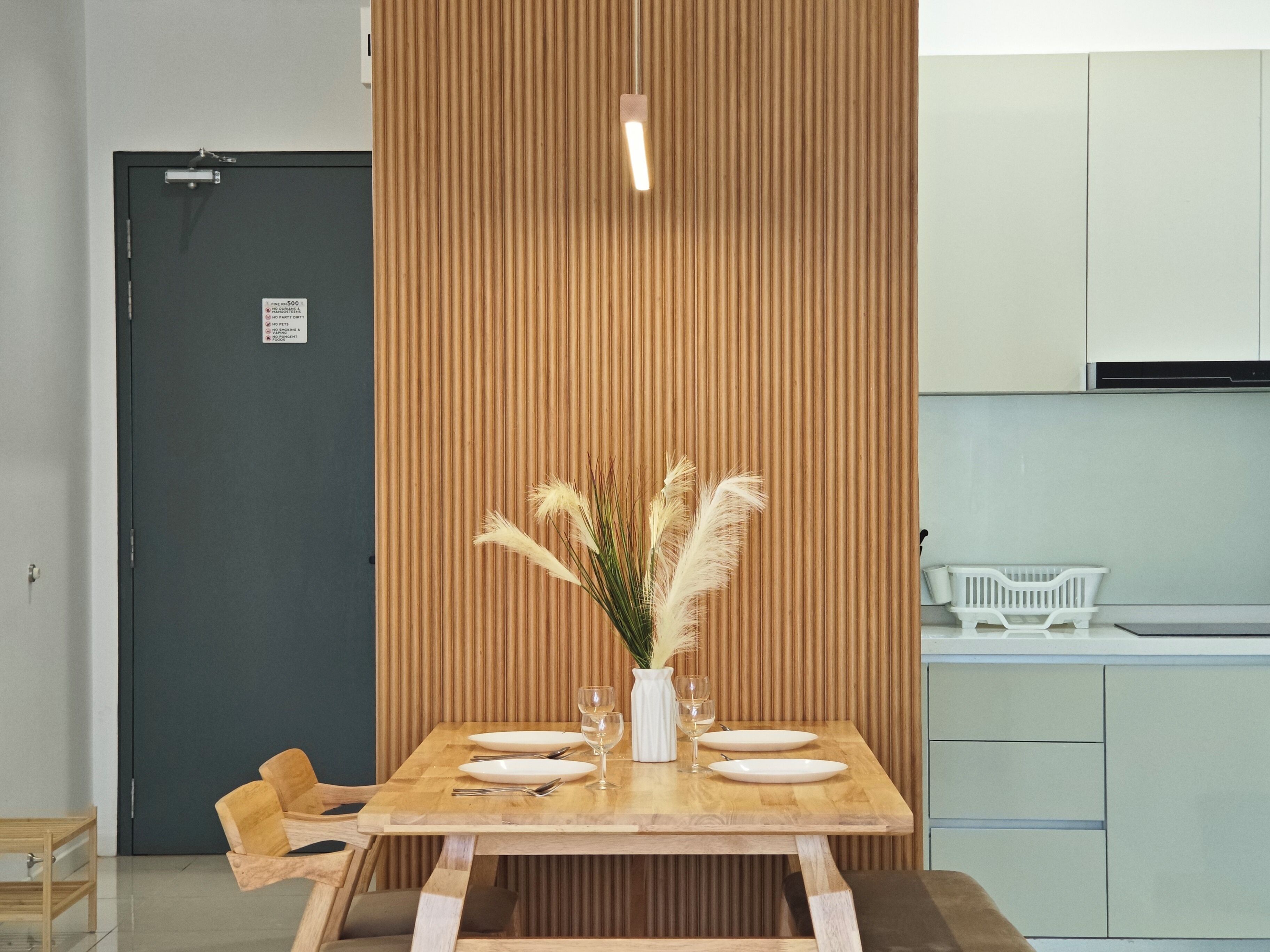 A dining area with a wooden table set for two, a vase with pampas grass, and a modern kitchen in the background.