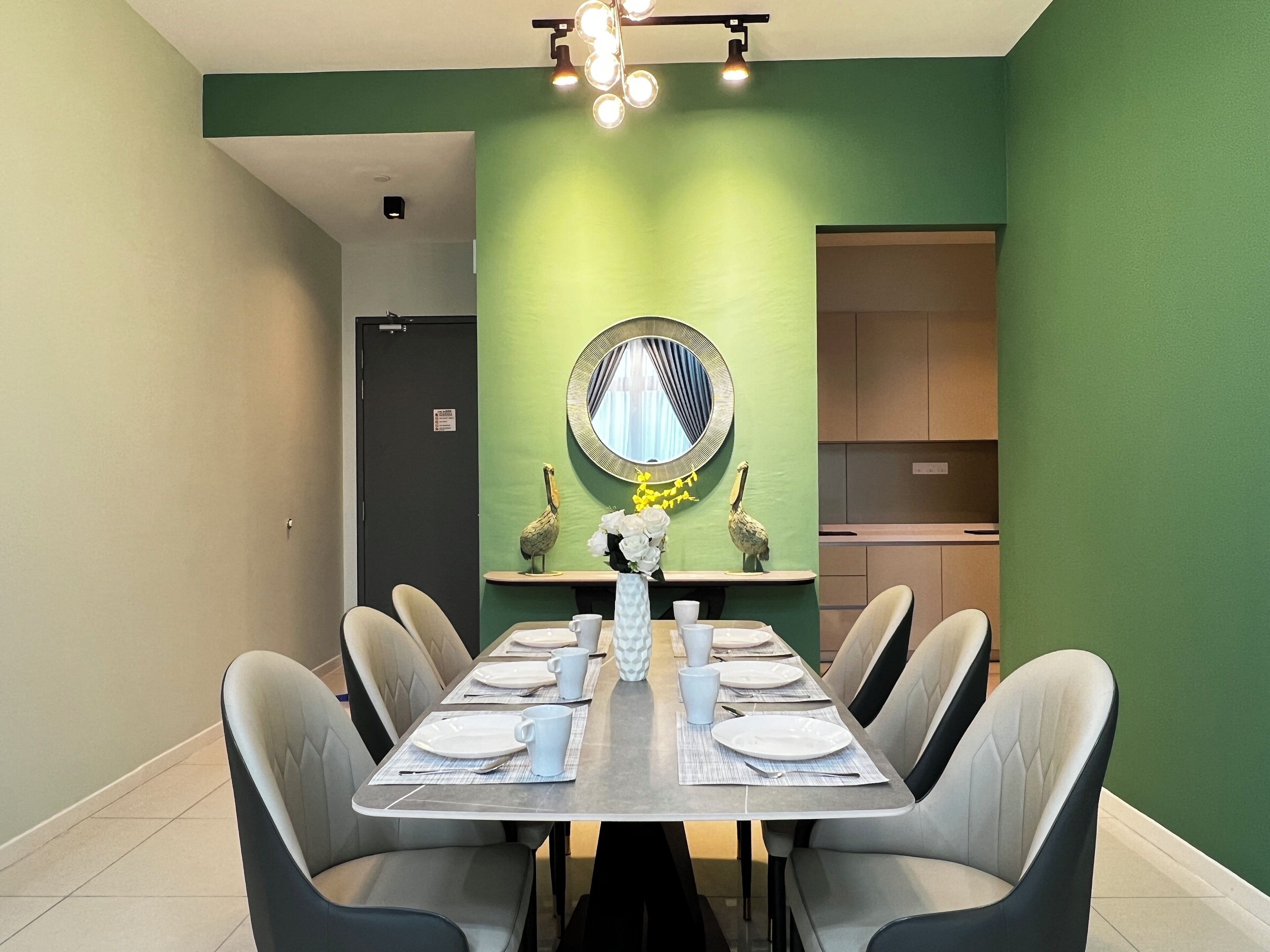 A dining area with a long table set for six, surrounded by chairs. A mirror with a decorative frame hangs on the green wall, and a small potted plant is on the table.
