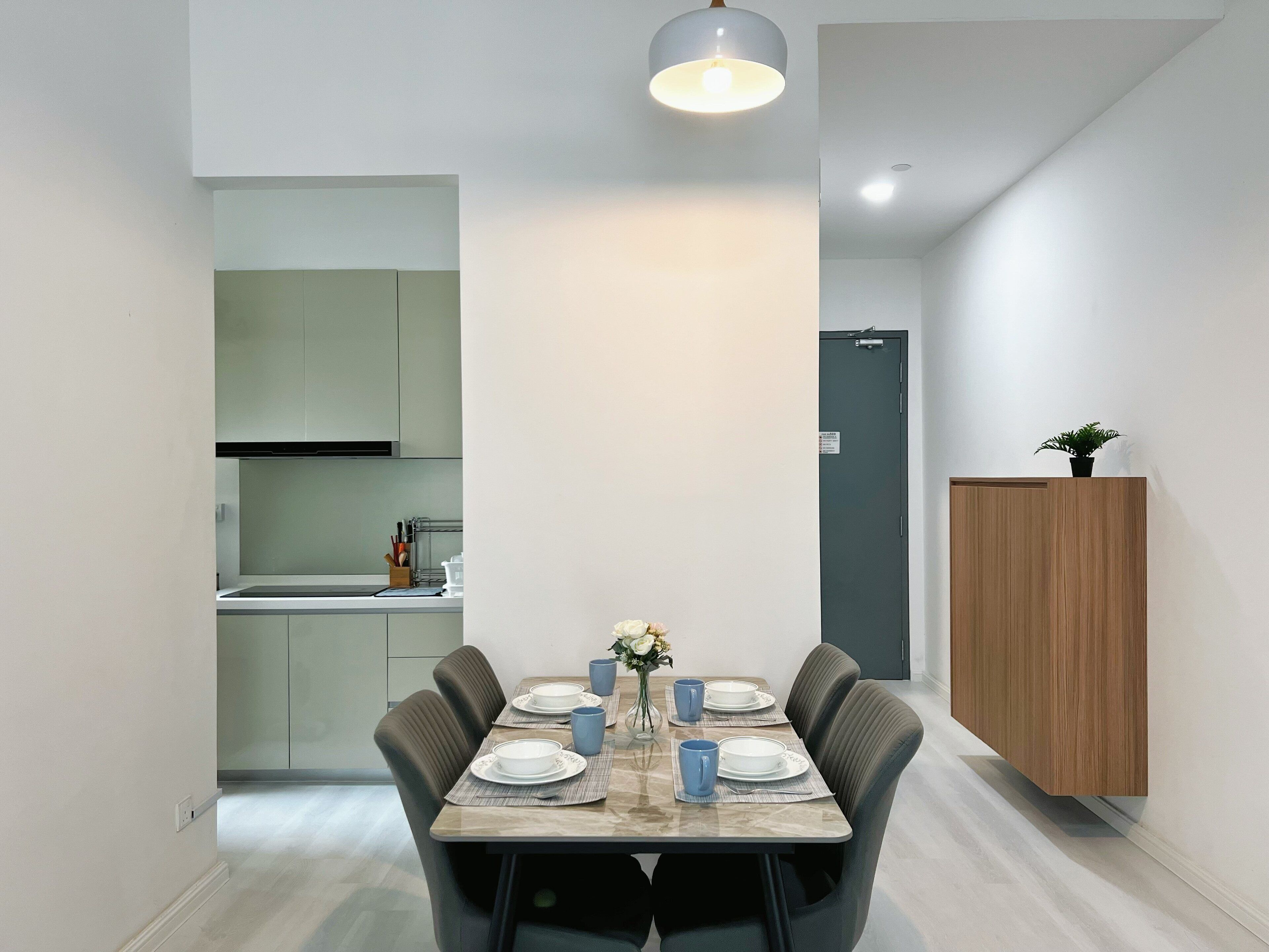A modern dining area with a rectangular table set for four, surrounded by dark chairs. A small plant is placed on a wooden cabinet to the right.