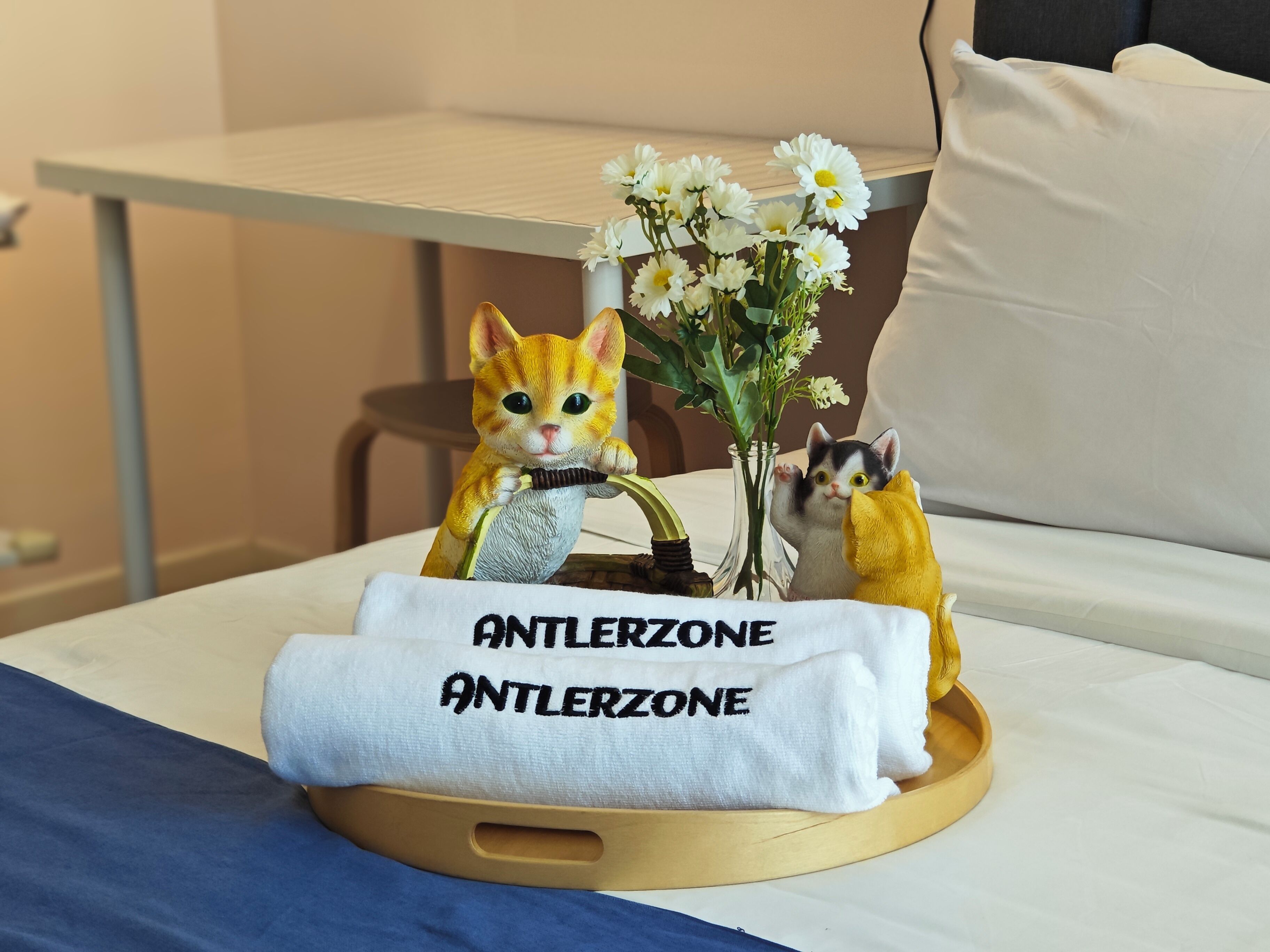 Una cama con una almohada blanca, una toalla blanca con la leyenda "ANTLERZONE" y un pequeño florero con flores blancas.