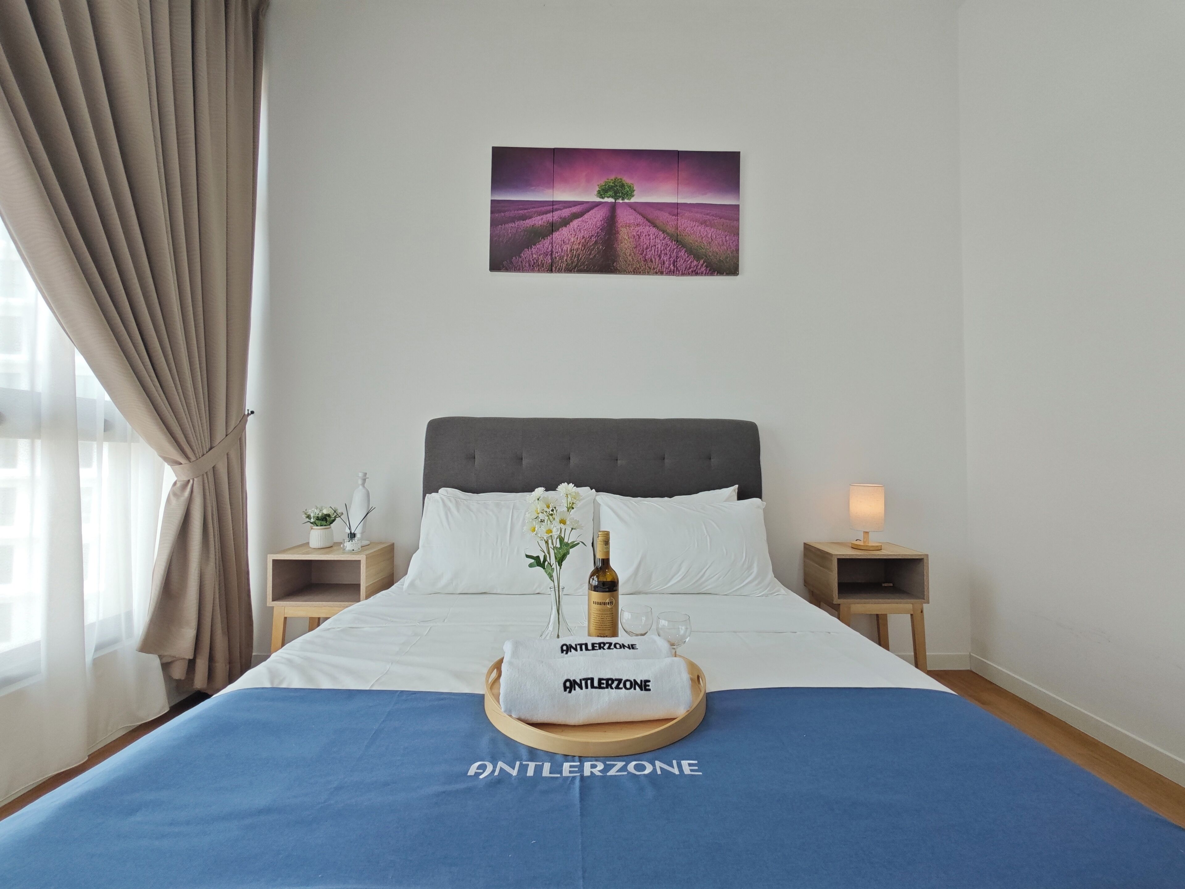 A hotel room with a bed, a nightstand with a bottle and glasses, a nightstand with a lamp, a window with curtains, and a painting of a purple field with a tree.