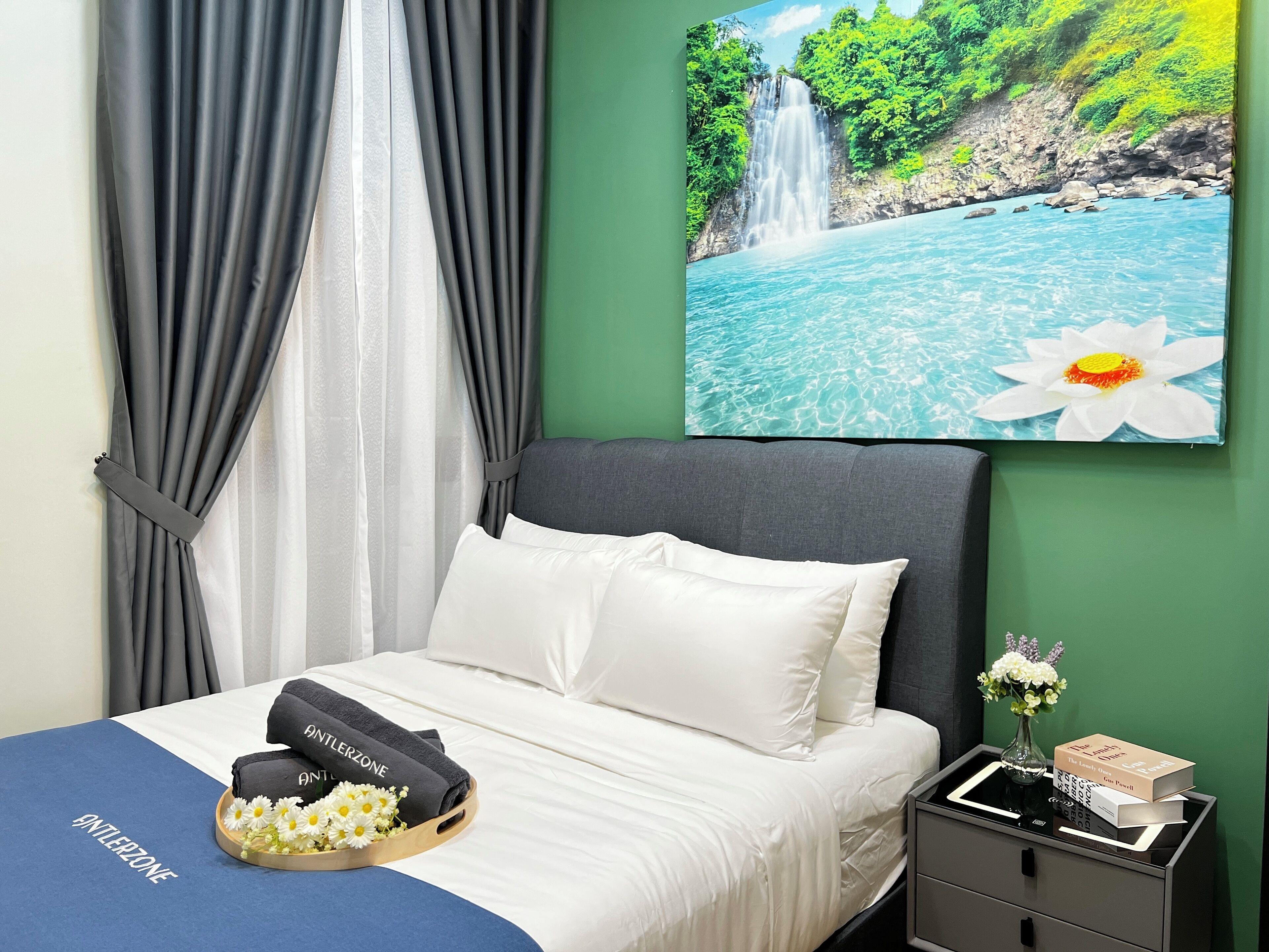 A bedroom with a bed, a nightstand, a painting of a waterfall, and a window with curtains.