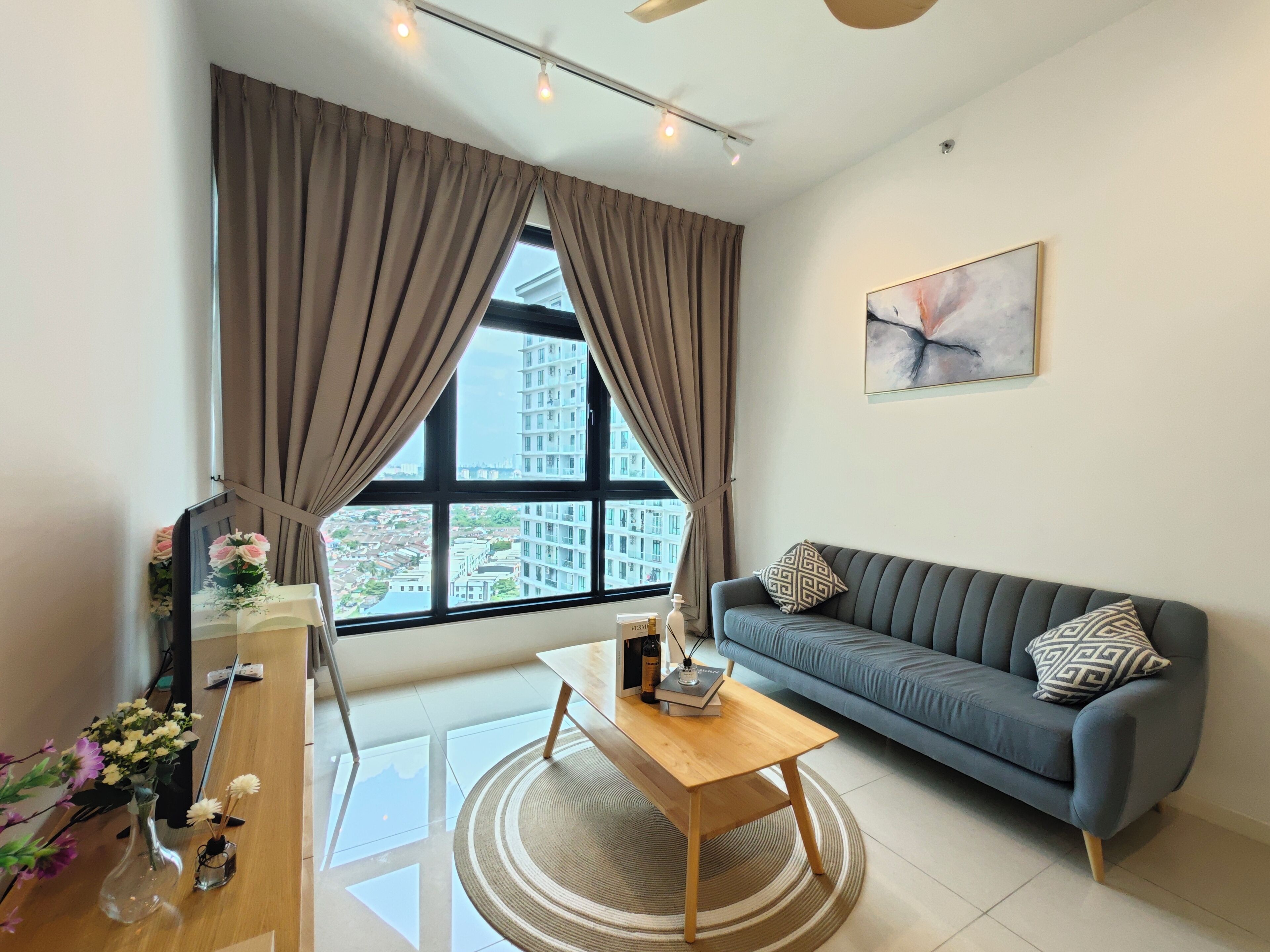 A modern living room with a sofa, a wooden coffee table, and a view of a cityscape through large windows with curtains.