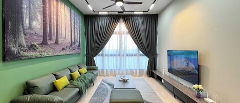 Una sala de estar moderna con un sofá verde, un reposapiés gris, un cuadro grande de un bosque, un ventilador de techo y un televisor sobre un mueble.