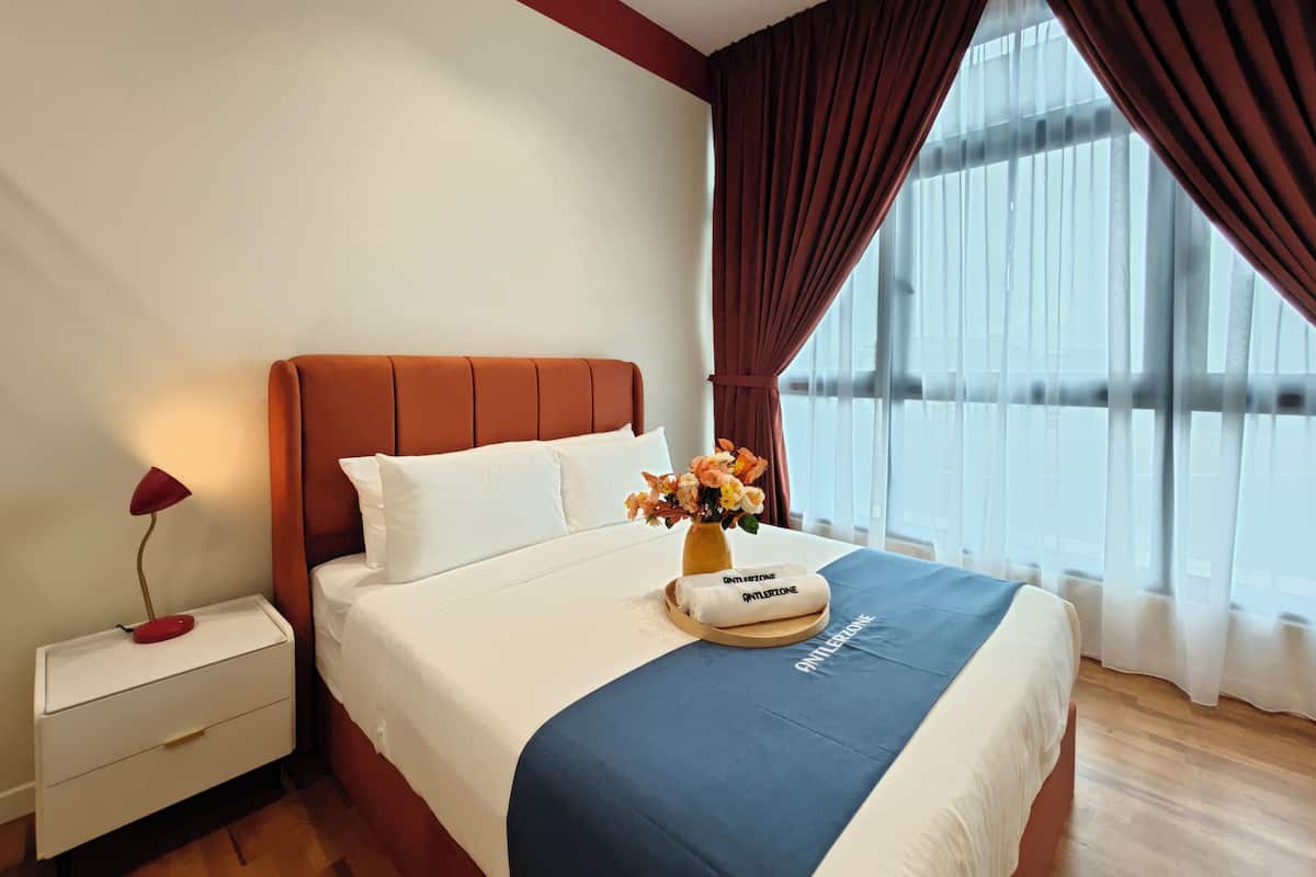Ein Hotelzimmer mit Bett, Nachttisch, Lampe, Blumen in einer Vase und einem Fenster mit Vorhängen.