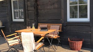 Ulkona oleva puinen terassi, jossa on pöytä ja tuoleja, rottinkikori ja ikkuna.