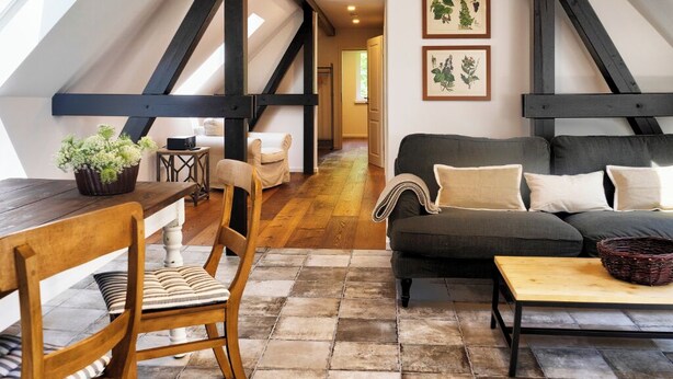 A cozy loft-style living space with a wooden dining table, a sofa, and a coffee table. A staircase leads to an upper level, and there are framed botanical prints on the wall.