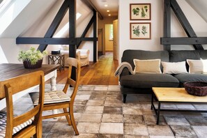 Un acogedor espacio tipo loft con una mesa de comedor de madera, un sofá y una mesita de centro. Una escalera conduce a un nivel superior, y hay impresiones botánicas enmarcadas en la pared.