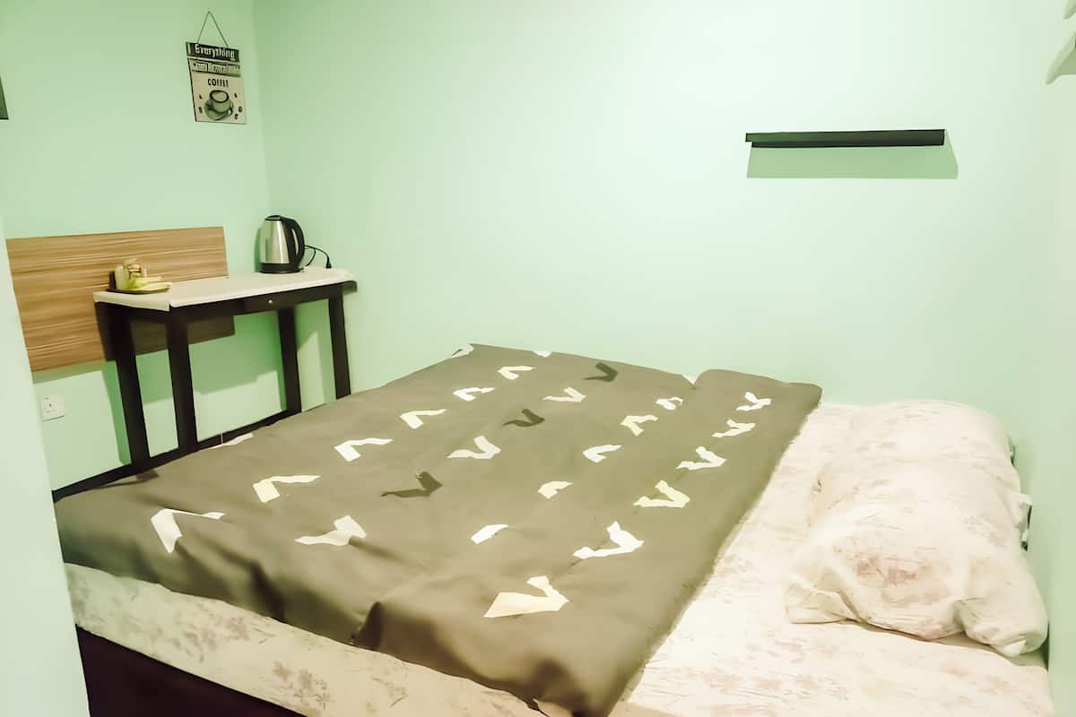 Ein kleiner, schlichter Raum mit einem Bett, einem Nachttisch, einem Wasserkocher und einer Wanduhr.
