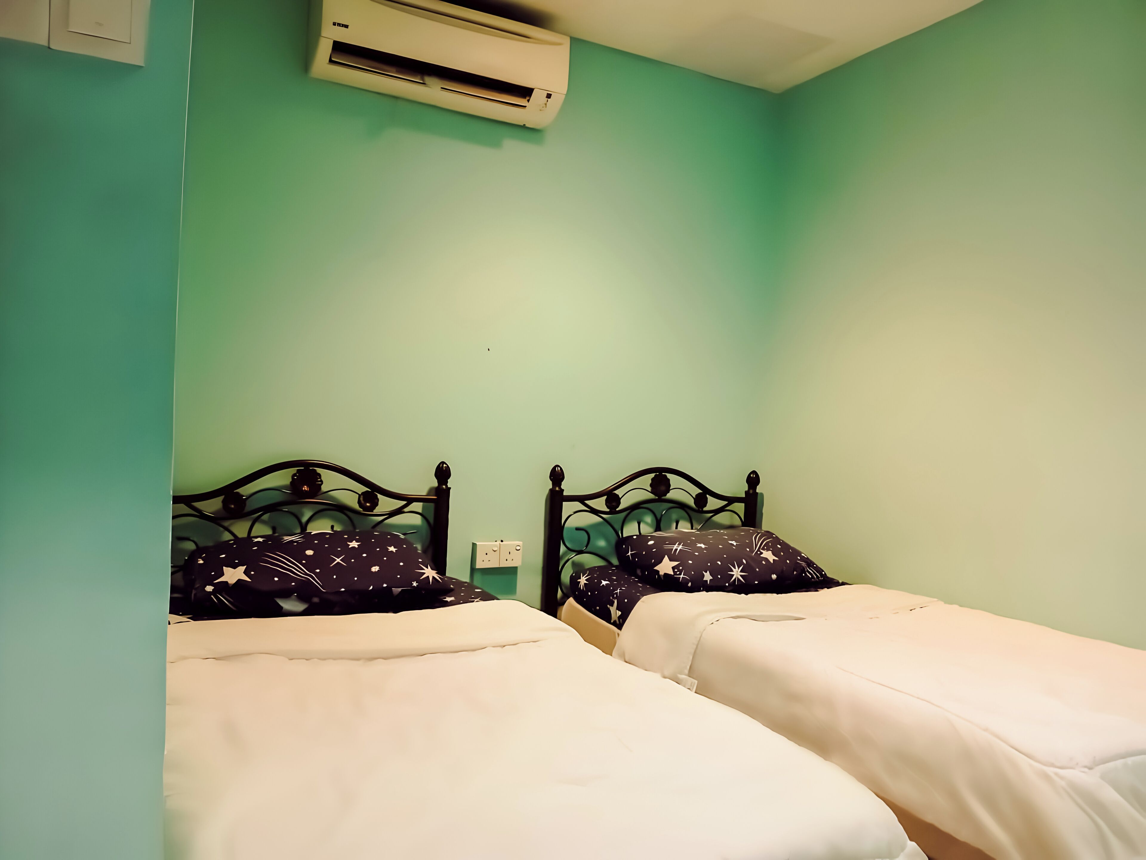 Two single beds with star-patterned pillows, a wall-mounted air conditioner, and a plain light switch.