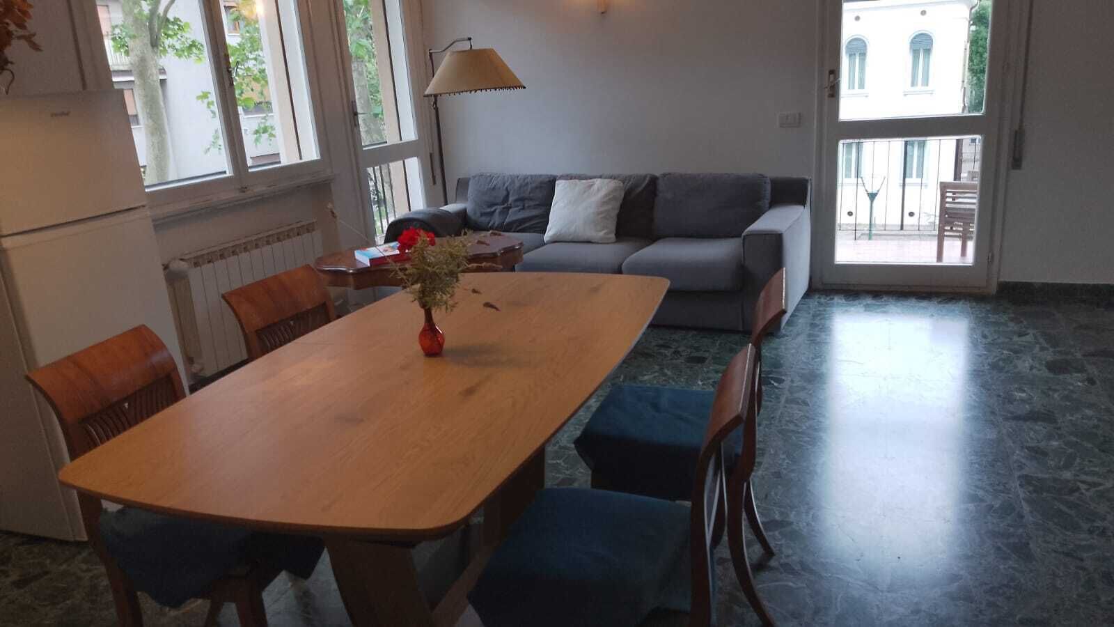 A dining area with a wooden table, chairs, and a vase of flowers. There is a sofa and a lamp in the background.