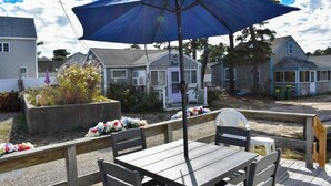 A patio with a table and chairs, a blue umbrella, and a wooden fence. - Completely renovated 3 bedroom cottage, .2 miles from Glendon Road beach (Dennis Port)