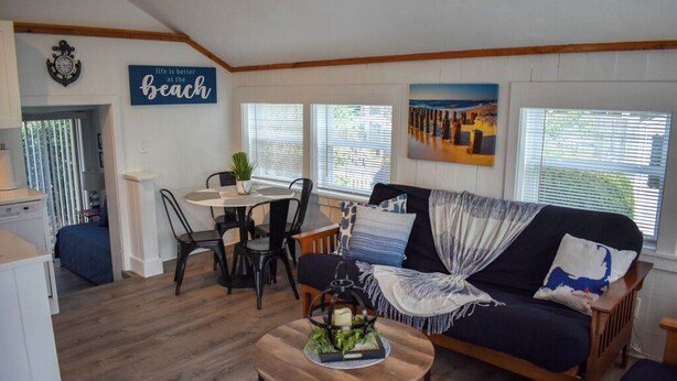 A cozy living room with a sofa, a coffee table, and a dining area with a table and chairs. There is a painting on the wall and a sign that says "life is better at the beach". - Completely renovated 3 bedroom cottage, .2 miles from Glendon Road beach (Dennis Port)
