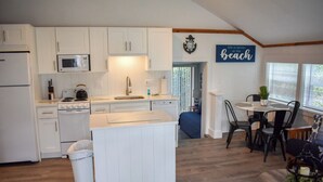 A modern kitchen with white cabinetry, a microwave, stove, and sink. There is a dining area with a table and chairs, and a blue accent wall with a 'Life is better at the beach' sign. - Completely renovated 3 bedroom cottage, .2 miles from Glendon Road beach (Dennis Port)